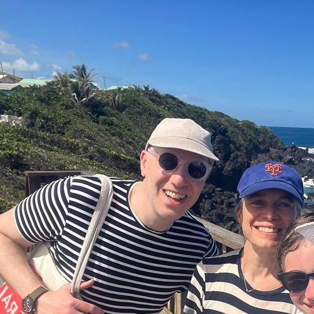 Three smiling guests in caps and striped shirts at a wooden railing, with a green hillside, dark volcanic rock and the ocean behind them