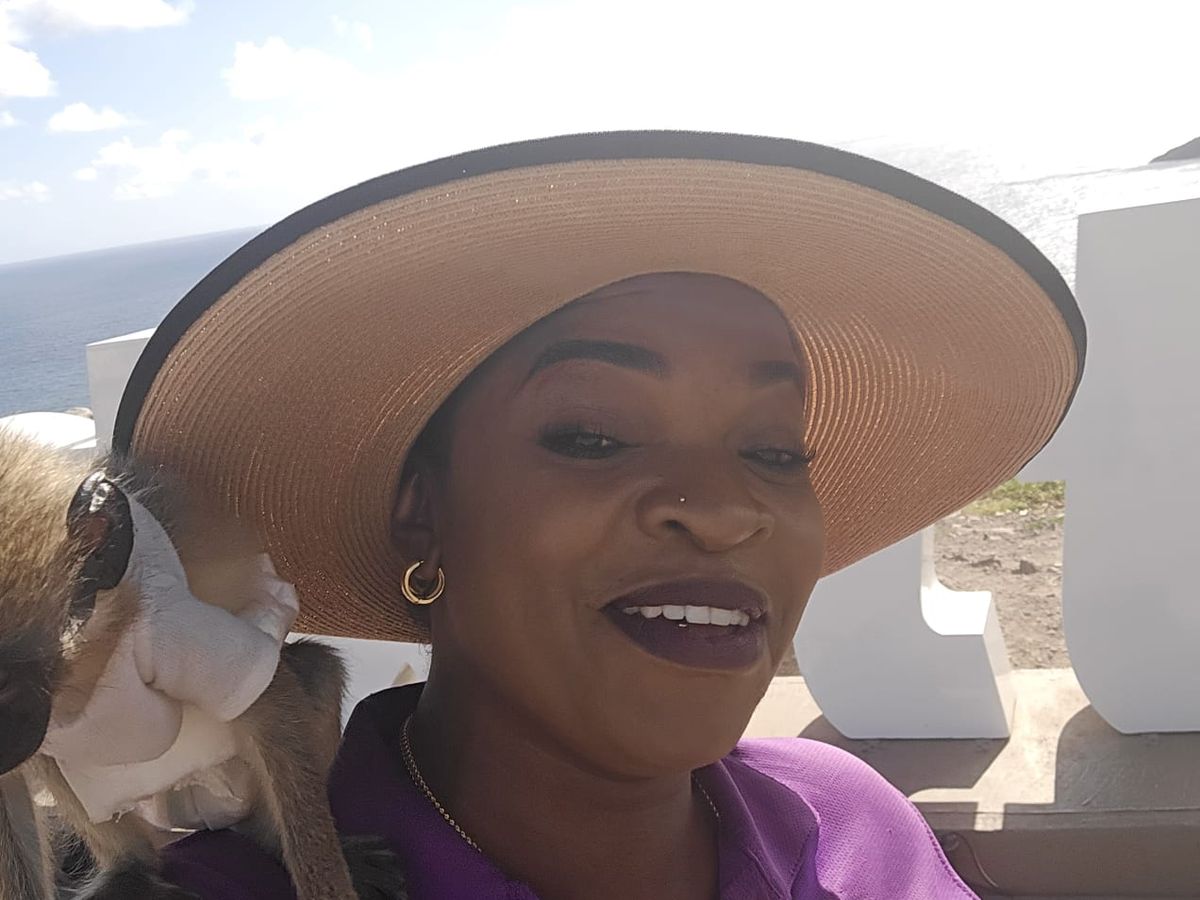 Mechiel Francis Dunrod in a wide sun hat and purple shirt, smiling, with a green vervet monkey sitting on her shoulder at a lookout with the sea behind her