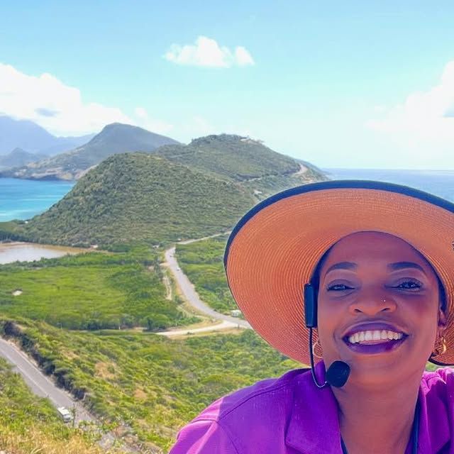 Mechiel in a sun hat and headset microphone at Timothy Hill on a sunny day, with the green hills of the Southeast Peninsula, its road and a salt pond behind her