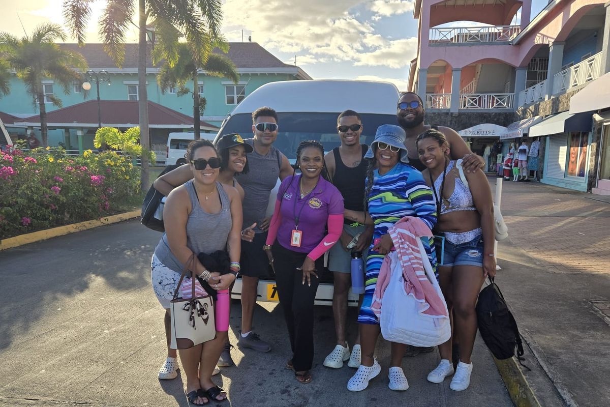 Mechiel in a purple shirt with a group of cruise guests in front of her white tour bus at Port Zante, Basseterre, with pink and blue buildings behind